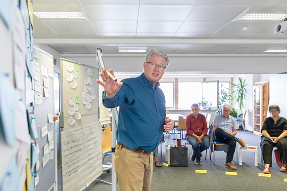 Ein Coach steht vor einem Whiteboard. Hinter ihm sitzen Teilnehmende des Vereinscoaching. 